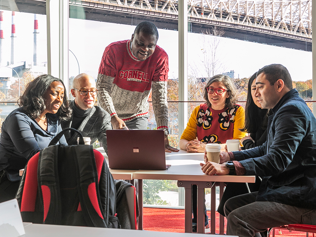 Students sit at a table gathered around a laptop on the Cornell Tech campus with the bridge to Roosevelt Island visible out the window.