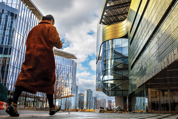 A person in an orange coat walks between two big buildings with a cityscape in the background.