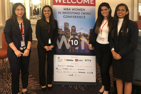 Four women in business attire standing around a Women In Investing Conference banner.