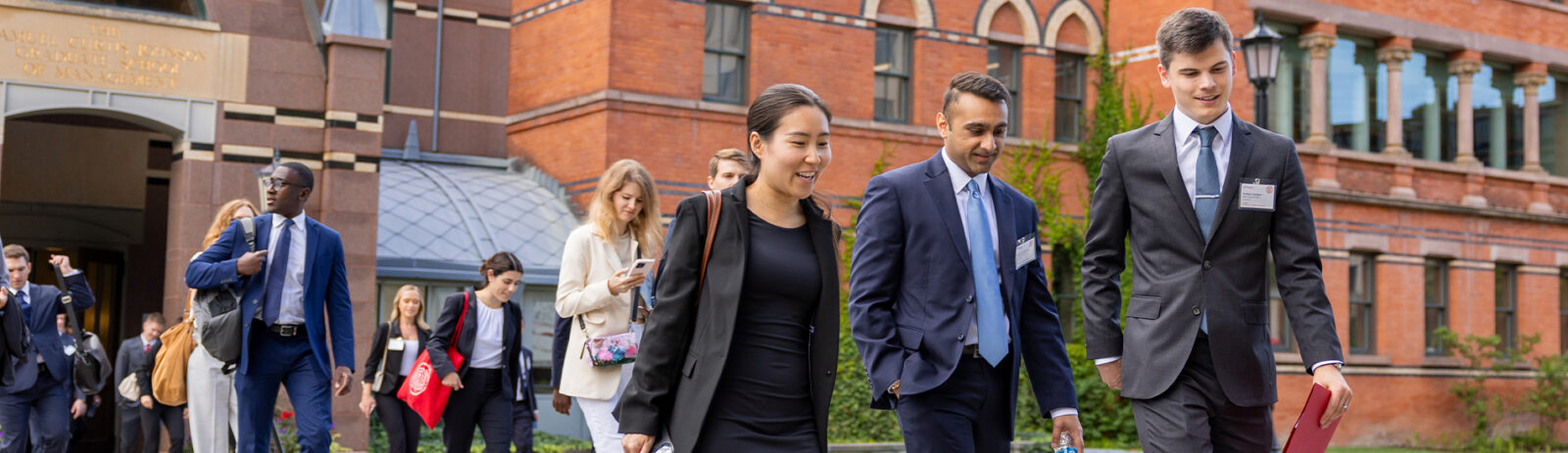 Students in business attire walk across the road with Sage Hall in the background.