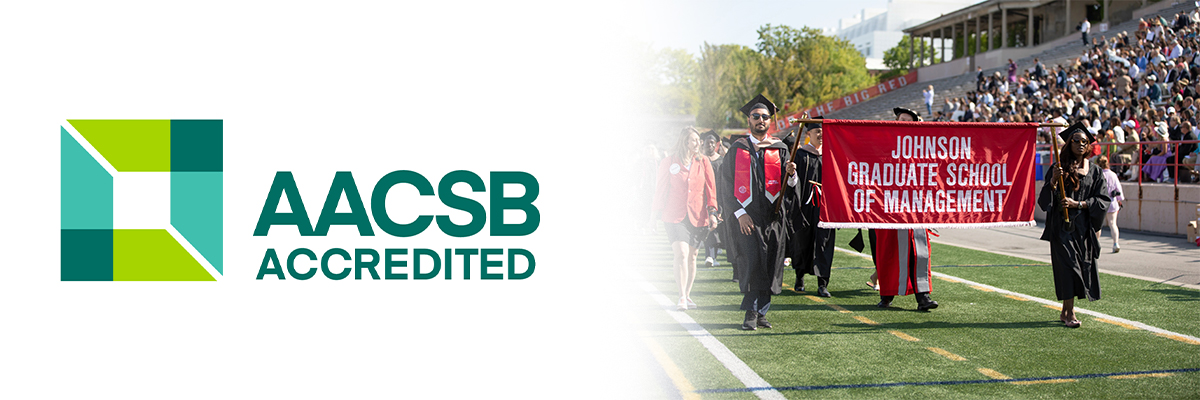 AACSB logo fading into a photo of students in graduation caps and gowns walking on a sports field holding a banner that says “Johnson Graduate School of Management.”