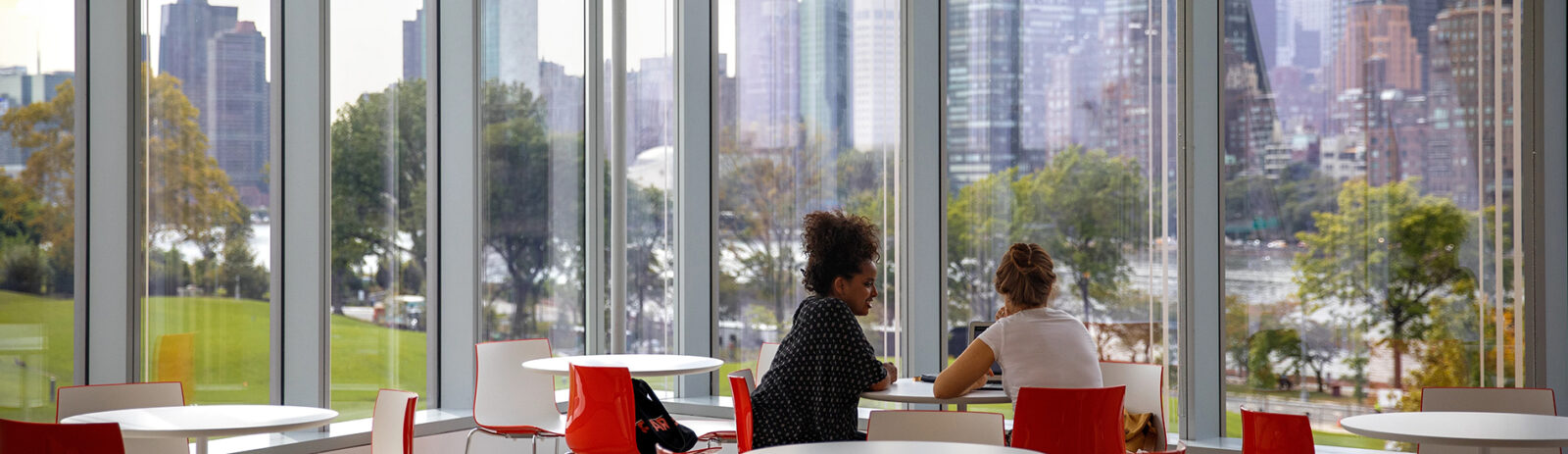 Two students sitting at a table with the city skyline seen through the window.
